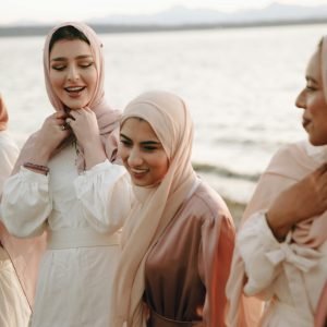 Four women in traditional hijabs sharing a joyful moment by the beach.